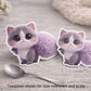 Two fluffy cat-shaped stickers next to a spoon on a textured surface.